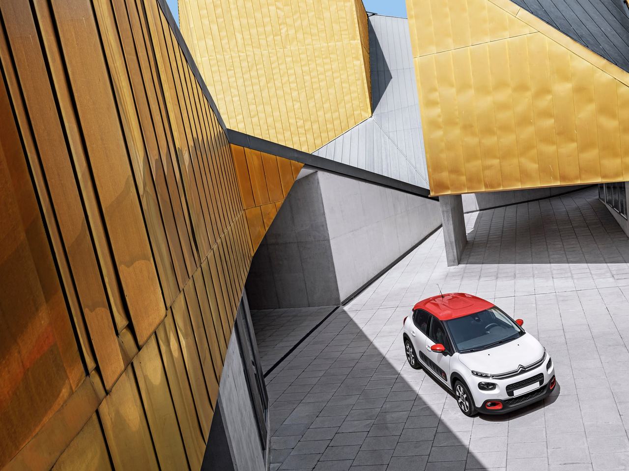 En vit Citroën C3 med rött tak ovanifrån, parkerad intill en gul fasad.